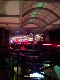 The image features the interior of a dimly lit bar or nightclub. The scene includes a bar with red neon lighting, various tables and chairs arranged in a cozy manner, and a decorative ceiling with arched beams. A disco ball hangs from the ceiling, adding to the glamorous atmosphere.
