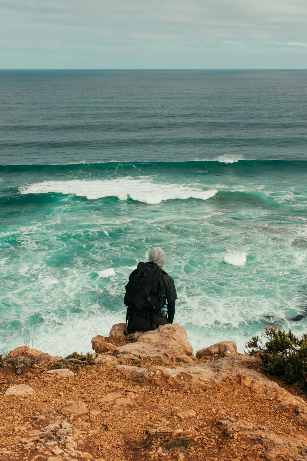 man in black jacket sitting on brown rock formation near body of water during daytime