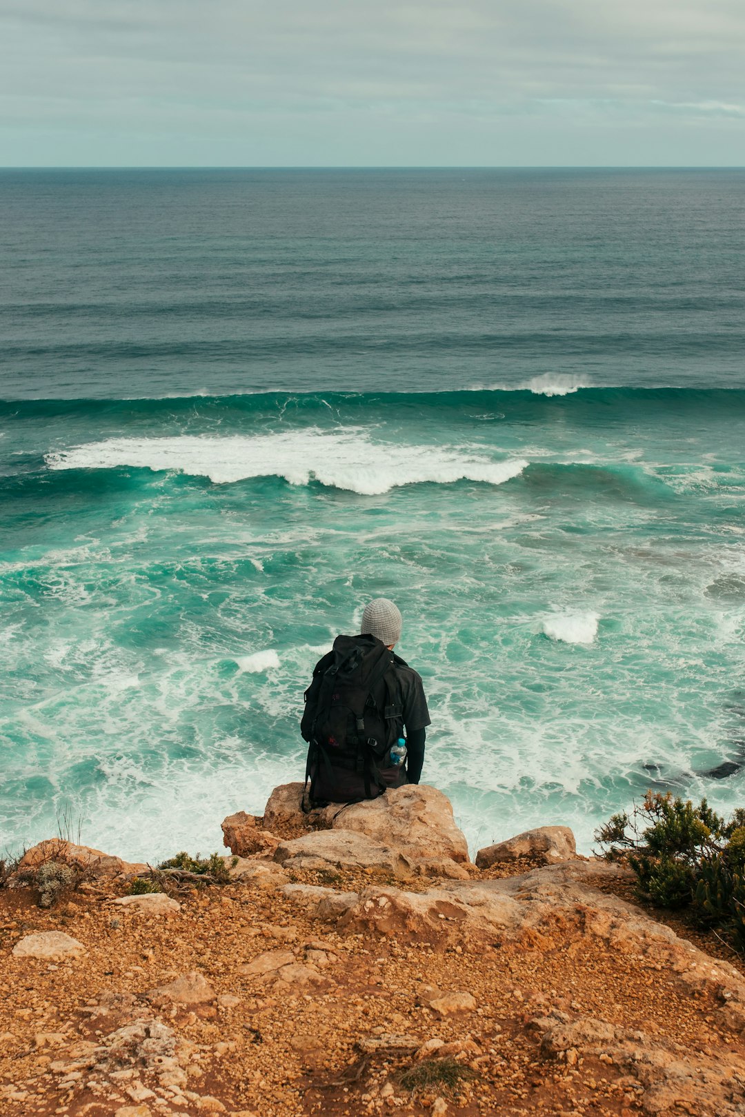 man in black jacket sitting on brown rock formation near body of water during daytime