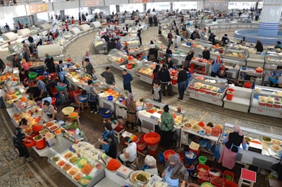 A bustling indoor market features numerous vendors selling a variety of fresh produce, meats, and prepared foods. Stalls are organized in a circular pattern, with sellers and customers interacting over counters lined with colorful fruits, vegetables, and containers. The atmosphere is lively with people moving around, examining goods, and engaging in commerce.