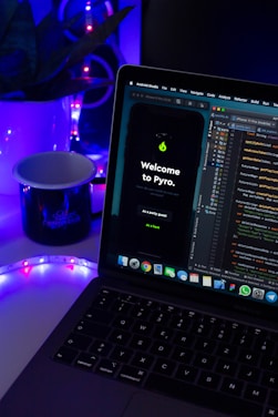 A laptop displaying code in an IDE is next to a mobile phone with an app featuring a green flame icon and the text 'Welcome to Pyro.' A white mug on the desk is illuminated by blue and purple ambient lighting. Potted plant leaves and a colorful LED strip add to the aesthetic setup.
