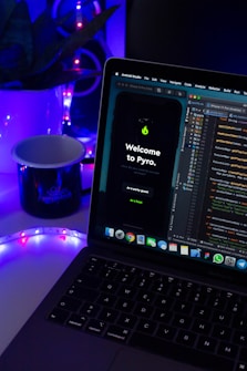 A laptop displaying code in an IDE is next to a mobile phone with an app featuring a green flame icon and the text 'Welcome to Pyro.' A white mug on the desk is illuminated by blue and purple ambient lighting. Potted plant leaves and a colorful LED strip add to the aesthetic setup.