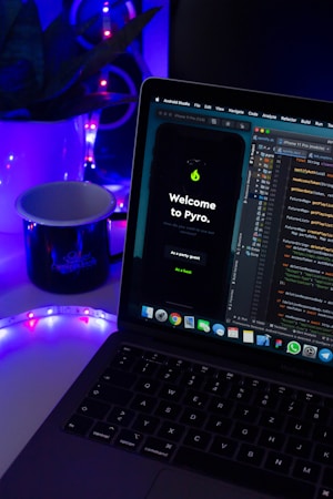 A laptop displaying code in an IDE is next to a mobile phone with an app featuring a green flame icon and the text 'Welcome to Pyro.' A white mug on the desk is illuminated by blue and purple ambient lighting. Potted plant leaves and a colorful LED strip add to the aesthetic setup.