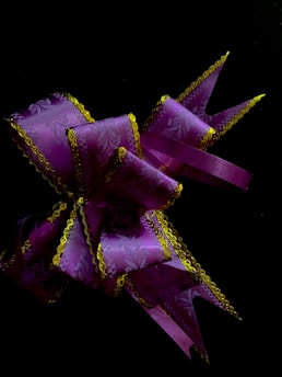 A decorative purple ribbon with intricate patterns and gold trim is elegantly arranged against a dark background. The ribbon features delicate floral designs and pointed, stylized ends.