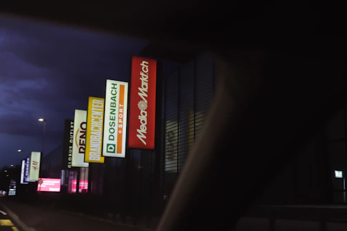 A vibrant mall strip featuring illuminated signs of global brand partners against a twilight sky.