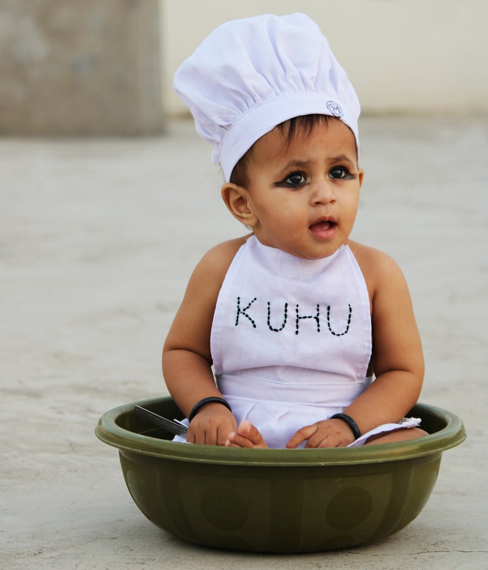 baby in white tank top and white cap sitting on gray plastic basin
