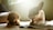 Two dogs are lying on a rug in a sunlit room, enjoying chew toys. One dog has curly, brown fur and is holding a toy, while the other has a smooth, light coat with darker patches and large ears.