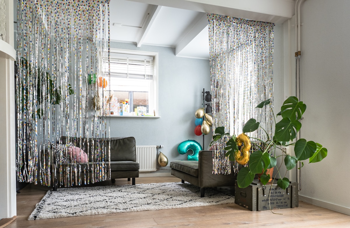 A simple living room with two grey couches in an L formation.  There is a white rug with a black large diamond patter.  The entrace is decorated with streaming tinsel curtains and there is a plant in front.  Random shiny metallic balloons are strewn around.  No one is in the room.