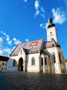 A historic church featuring a colorful tiled roof with intricate patterns and symbols stands in a cobblestone plaza under a bright blue sky. Its tall bell tower is capped with a dark spire, and the building's facade is adorned with arched windows and detailed stonework.