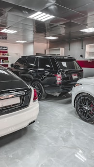 A showroom or garage setting featuring three luxury vehicles closely parked. The cars include a black SUV, a white car, and another white vehicle partially visible, with glossy finishes and polished appearances. The environment is well-lit with reflective ceiling panels and fluorescent lights casting reflections on the cars.