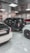 A showroom or garage setting featuring three luxury vehicles closely parked. The cars include a black SUV, a white car, and another white vehicle partially visible, with glossy finishes and polished appearances. The environment is well-lit with reflective ceiling panels and fluorescent lights casting reflections on the cars.