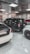 A showroom or garage setting featuring three luxury vehicles closely parked. The cars include a black SUV, a white car, and another white vehicle partially visible, with glossy finishes and polished appearances. The environment is well-lit with reflective ceiling panels and fluorescent lights casting reflections on the cars.
