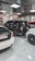 A showroom or garage setting featuring three luxury vehicles closely parked. The cars include a black SUV, a white car, and another white vehicle partially visible, with glossy finishes and polished appearances. The environment is well-lit with reflective ceiling panels and fluorescent lights casting reflections on the cars.