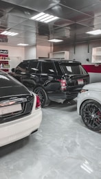 A showroom or garage setting featuring three luxury vehicles closely parked. The cars include a black SUV, a white car, and another white vehicle partially visible, with glossy finishes and polished appearances. The environment is well-lit with reflective ceiling panels and fluorescent lights casting reflections on the cars.