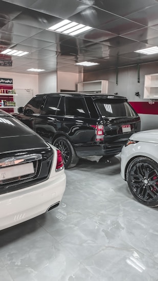 A showroom or garage setting featuring three luxury vehicles closely parked. The cars include a black SUV, a white car, and another white vehicle partially visible, with glossy finishes and polished appearances. The environment is well-lit with reflective ceiling panels and fluorescent lights casting reflections on the cars.