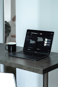 A laptop with a dark screen featuring a software interface is placed on a dark wooden table. Next to the laptop is a black mug, and in the background, there is a softly blurred view of a room with light walls.