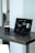 A laptop with a dark screen featuring a software interface is placed on a dark wooden table. Next to the laptop is a black mug, and in the background, there is a softly blurred view of a room with light walls.