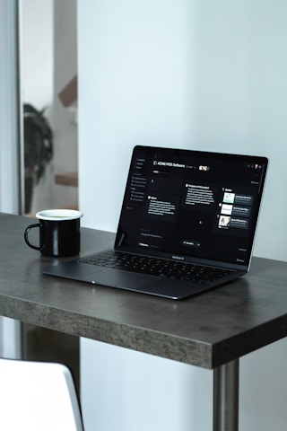 A laptop with a dark screen featuring a software interface is placed on a dark wooden table. Next to the laptop is a black mug, and in the background, there is a softly blurred view of a room with light walls.
