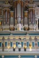 An ornate and intricately designed pipe organ with golden decorative elements and blue-gray panels dominates the scene. Below the organ pipes, a person is seated, partially visible from behind, possibly playing the organ.