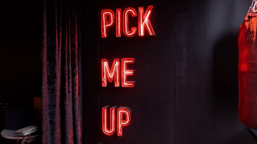 Neon letters in bright red spell out the words 'PICK ME UP' vertically against a dark background. To the right, part of a red leather punching bag is visible, while a dark curtain hangs on the left.