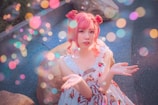 A person with pink hair styled in two buns is wearing a frilly, floral dress. They are sitting or crouching near large rocks on a surface that appears to be blue pavement. The image features numerous multicolored bokeh light effects, adding a whimsical and dreamy atmosphere.