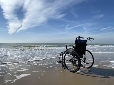 A wheelchair is positioned on a sandy beach at the edge of the water, with gentle waves lapping at the shore. The sky is mostly clear with some wispy clouds, creating a serene and peaceful coastal scene.