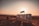 Large white letters spelling 'AGADIR' are situated on a sandy beach with the sun setting in the background. The sky is tinged with warm hues of orange and pink. A few people can be seen walking along the shore, and the ocean appears calm.