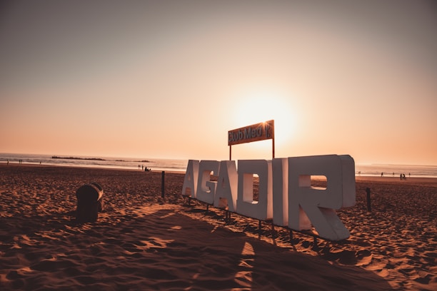 Large white letters spelling 'AGADIR' are situated on a sandy beach with the sun setting in the background. The sky is tinged with warm hues of orange and pink. A few people can be seen walking along the shore, and the ocean appears calm.