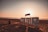 Large white letters spelling 'AGADIR' are situated on a sandy beach with the sun setting in the background. The sky is tinged with warm hues of orange and pink. A few people can be seen walking along the shore, and the ocean appears calm.