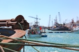 A nautical scene featuring a close-up of a rusty anchor with two ropes tied to it, casting a vivid contrast against the turquoise sea in the background. Various boats are moored at a harbor, with cranes and industrial structures visible, suggesting a working port. The sky is clear and blue, enhancing the overall brightness.