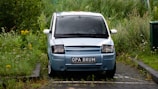A blue compact car is parked on a small paved driveway surrounded by overgrown grass and wild plants. The car has a distinctive registration plate displayed at the front. The surrounding area appears to be a neglected or less maintained parking space with greenery encroaching onto the pavement.