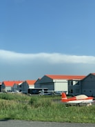 Several small aircraft are parked in front of a row of hangars with orange roofs. The hangars are situated in a grassy area under a clear blue sky.