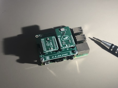 A small electronic board with multiple ports and labeled components, sitting on a plain surface under direct light. An electronic screwdriver is positioned to the right, casting a shadow next to the board.