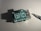 A small electronic board with multiple ports and labeled components, sitting on a plain surface under direct light. An electronic screwdriver is positioned to the right, casting a shadow next to the board.