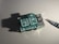 A small electronic board with multiple ports and labeled components, sitting on a plain surface under direct light. An electronic screwdriver is positioned to the right, casting a shadow next to the board.