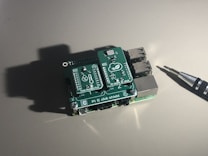 A small electronic board with multiple ports and labeled components, sitting on a plain surface under direct light. An electronic screwdriver is positioned to the right, casting a shadow next to the board.