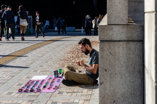 A bearded man sits on the ground next to a stone wall, engaged in crafting or arranging objects. In front of him lies a purple mat displaying numerous colorful handmade items, possibly bracelets or small decorations. Beside the mat, there are several cans possibly containing paints or materials. In the background, people walk by on a paved surface, suggesting an outdoor public space or market.