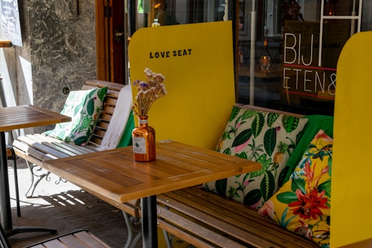 Outdoor seating arrangement with a wooden table and bench adorned with vibrant, tropical-themed cushions. A tall, yellow divider is marked with the words 'LOVE SEAT', providing a cozy and inviting atmosphere. A small glass bottle containing dried flowers serves as a centerpiece on the table, adding a rustic touch. The background includes a store window with the text 'BIJ ETEN&'.