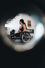 A person with long hair is performing a deadlift in an indoor gym setting. They are holding a barbell with weights labeled 'REP FITNESS'. The room is spacious with high ceilings and has posters on the walls. Another person is visible in the background, and the scene is captured through a circular vignette effect.