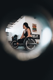 A person with long hair is performing a deadlift in an indoor gym setting. They are holding a barbell with weights labeled 'REP FITNESS'. The room is spacious with high ceilings and has posters on the walls. Another person is visible in the background, and the scene is captured through a circular vignette effect.