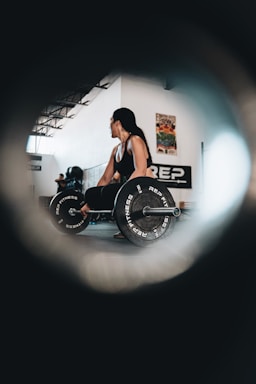 A person with long hair is performing a deadlift in an indoor gym setting. They are holding a barbell with weights labeled 'REP FITNESS'. The room is spacious with high ceilings and has posters on the walls. Another person is visible in the background, and the scene is captured through a circular vignette effect.