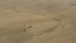 An aerial view of a vast farmland with a tractor attached to a large piece of agricultural machinery, moving across the field. The landscape is mostly flat and covered with brown earth, indicating either freshly plowed or harvested land.