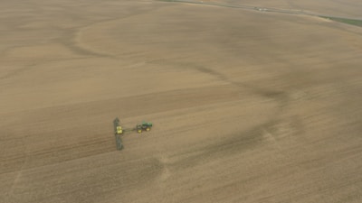 An aerial view of a vast farmland with a tractor attached to a large piece of agricultural machinery, moving across the field. The landscape is mostly flat and covered with brown earth, indicating either freshly plowed or harvested land.
