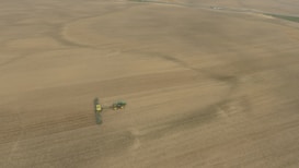 An aerial view of a vast farmland with a tractor attached to a large piece of agricultural machinery, moving across the field. The landscape is mostly flat and covered with brown earth, indicating either freshly plowed or harvested land.