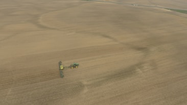 An aerial view of a vast farmland with a tractor attached to a large piece of agricultural machinery, moving across the field. The landscape is mostly flat and covered with brown earth, indicating either freshly plowed or harvested land.