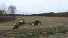 A green tractor with yellow wheels is towing a farming implement across a large, open field. The terrain is mostly flat with some residual crop stubble, bordered by a line of trees in the background. The scene appears overcast, with a cloudy sky.