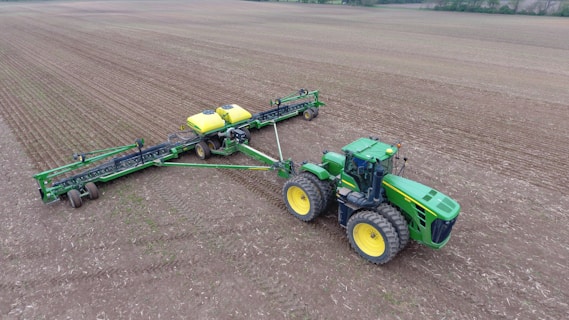 A large green tractor with yellow wheels is attached to a wide farming implement on a vast, tilled field. The machinery appears to be used for planting or seeding, evident from the evenly spaced rows in the soil. The landscape is expansive and flat, with a line of trees in the background under an overcast sky.