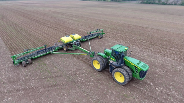 A large green tractor with yellow wheels is attached to a wide farming implement on a vast, tilled field. The machinery appears to be used for planting or seeding, evident from the evenly spaced rows in the soil. The landscape is expansive and flat, with a line of trees in the background under an overcast sky.