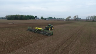 A large agricultural field with a tractor and planter in the center, preparing the ground. The scene is expansive with flat, recently tilled soil, bordered on the horizon by trees and a few farm buildings. The sky is overcast, lending a muted mood to the scene.