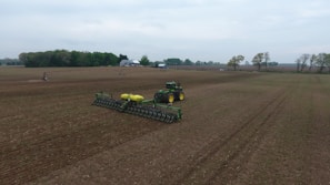A large agricultural field with a tractor and planter in the center, preparing the ground. The scene is expansive with flat, recently tilled soil, bordered on the horizon by trees and a few farm buildings. The sky is overcast, lending a muted mood to the scene.